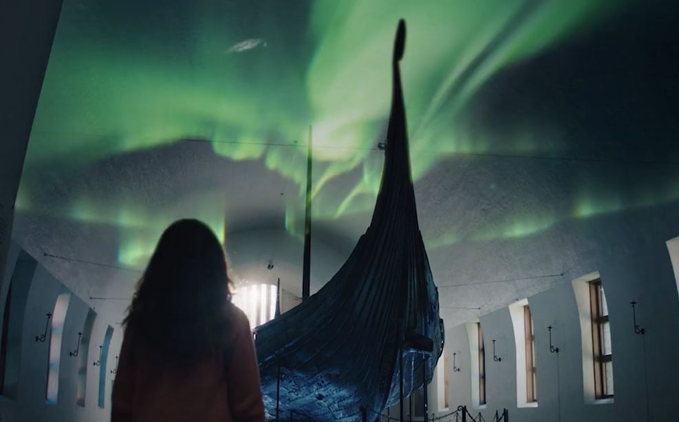 Jente med langt hår står og ser på et langskip med nordlyset i taket over. Foto/illustrasjon.