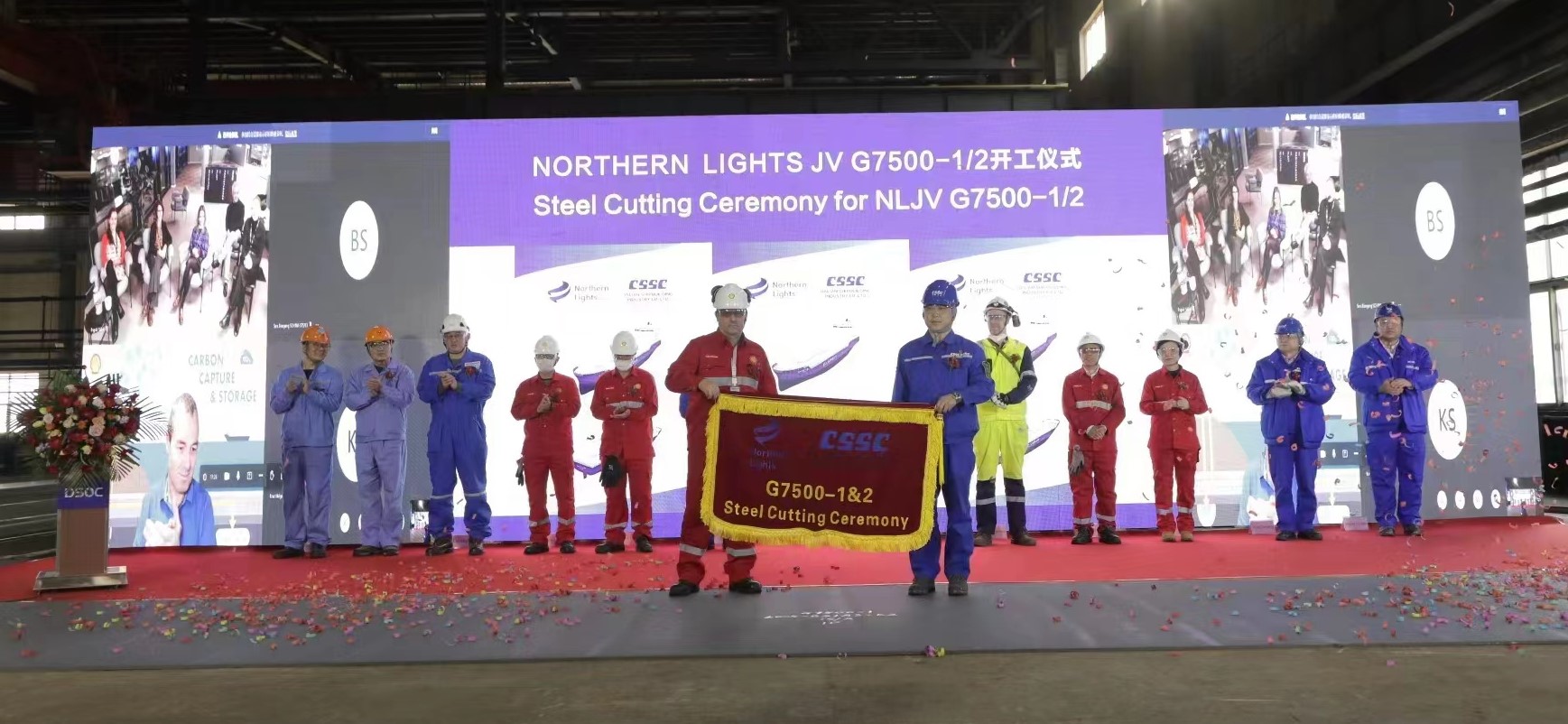 Flere mennesker i arbeidsklær og industrihjelmer som holder opp et banner med teksten "Steel Cutting Ceremony". Foto.