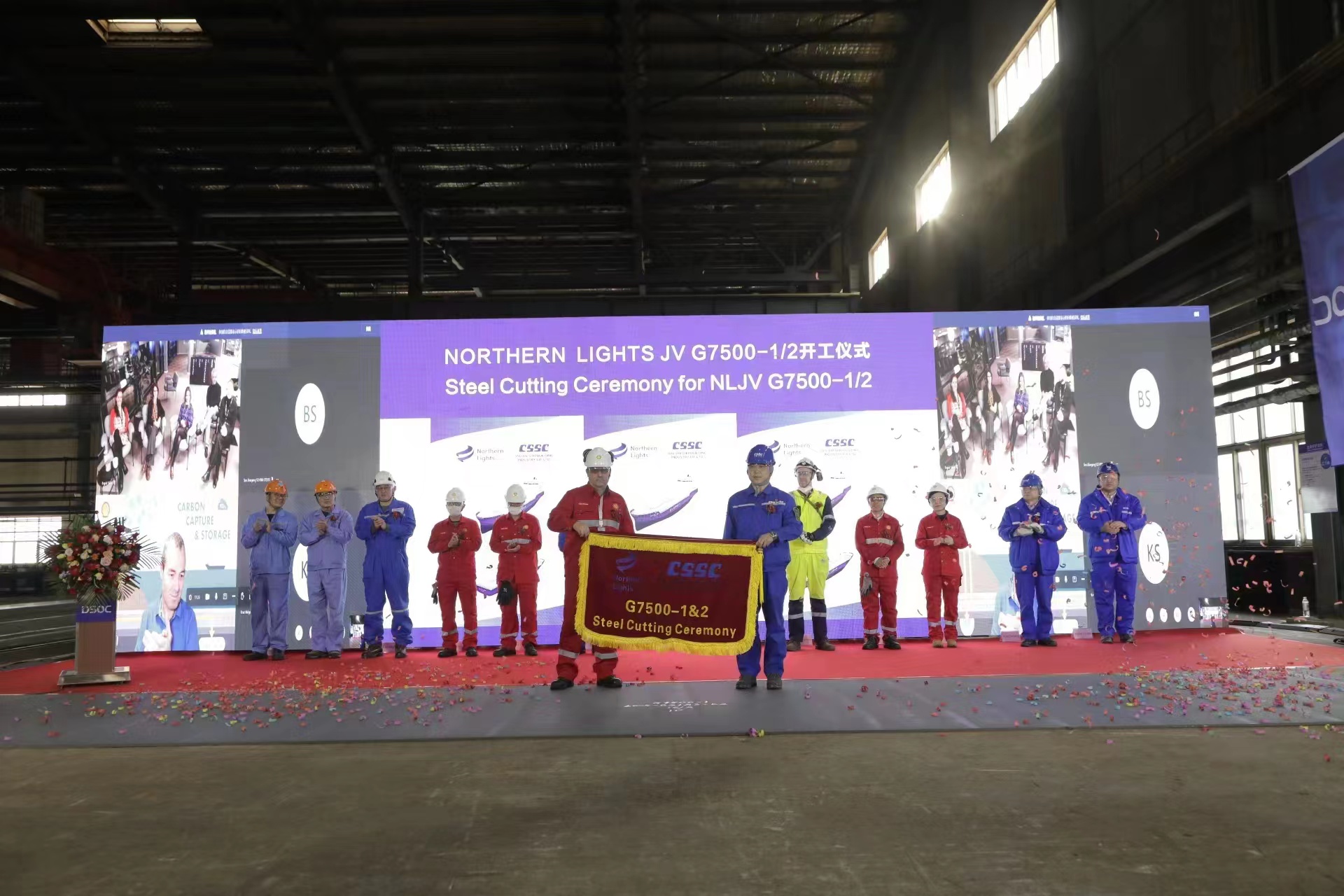 Flere mennesker i arbeidsklær og industrihjelmer som holder opp et banner med teksten "Steel Cutting Ceremony". Foto.